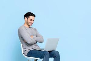 Portrait of smiling happy young Asian man sitting and looking at laptop computer with arms crossed isolated on light blue studio background photo