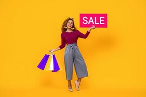 Smiling beautiful African American woman with colorful  shopping bags showing red sale sign standing on isolated yellow background photo