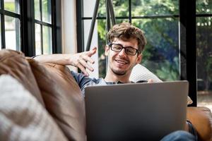 joven caucásico feliz chateando en línea a través de una videollamada con una computadora portátil en el sofá de casa