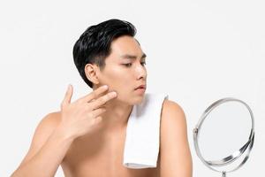 Shirtless young handsome Asian man looking at his face in the mirror  isolated on white background for skin care and beauty concepts