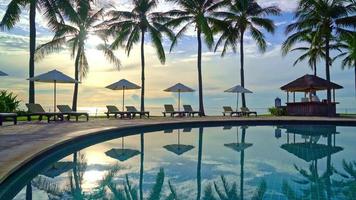 hermosa sombrilla y silla de lujo alrededor de la piscina al aire libre con palmeras de coco en el cielo del atardecer o del amanecer - concepto de vacaciones y vacaciones video