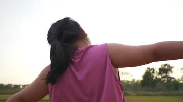 Back view of happy Asian fat girl playing in field, enjoy on holiday, standing on grass in the big park with a lot of trees and sunlight. Beautiful nature concept video