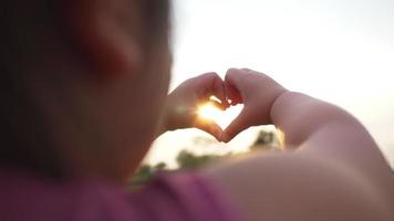 närbild händer på vacker flicka som gör hjärtform med händer i solnedgången flicka håller upp kärlekssymbolen i en vacker utsikt, grönt fält med solljuskoncept video