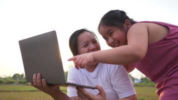 Close up of Asian fat girl and mom using laptop in field , enjoy on holiday, standing and playing laptop on grass in the big park with a lot of trees and sunlight