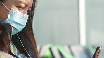 Close up Asian woman sitting and taking video call on smartphone at airport. Young woman wearing mask and waiting for airplane taking off at the airport. Using smartphone and earphone