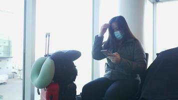 aziatische vrouw die een masker draagt, zit en smartphone gebruikt op de luchthaven. wachten op de vlucht op de luchthaven met haar koffer. met het vliegtuig naar huis op vakantie video