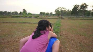 Rückansicht des asiatischen Mädchens und Vaters, die auf dem Feld spielen, Tochter reitet auf dem Rücken des Vaters auf Gras im großen Park mit vielen Bäumen im Hintergrund. schöne natur im abendkonzept