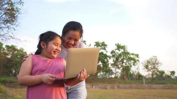 menina gorda asiática e mãe usando laptop em campo, aproveite as férias, em pé e jogando laptop na grama no grande parque com muitas árvores. bela natureza no conceito de noite video