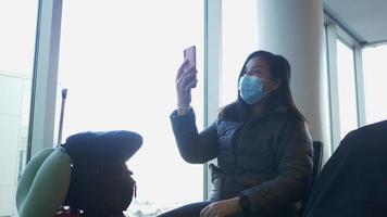 Asian woman sitting and taking video call on smartphone at airport. Young woman wearing mask and waiting for airplane taking off at the airport. Using smartphone and earphone