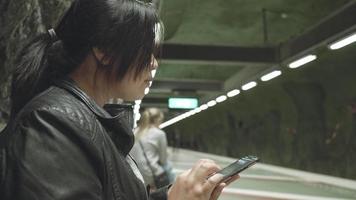 mujer asiática de pie, usando un teléfono inteligente y esperando el tren para llegar a casa. tomar el transporte público para llegar a casa. viajar solo al extranjero video