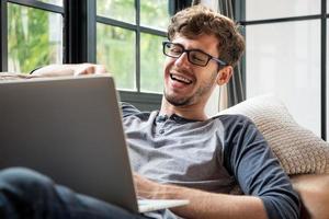 joven caucásico feliz riéndose mientras hace una videollamada chateando en línea con una computadora portátil en el sofá de casa foto