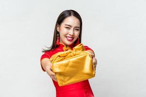 Portrait of smiling Asian woman in red traditional dress costume giving gold gift box in light gray isolated studio background for Chinese new year concepts photo