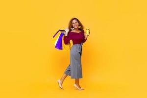 Full length portrait of cheerful young smiling shopaholic African American woman walking while holding shopping bags and drink cup in isolated studio yellow background
