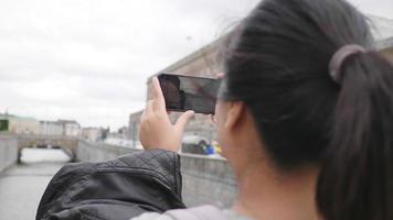 rückansicht einer asiatischen frau, die steht und ein foto von fluss und stadt in schweden macht, im urlaub ins ausland reist. auf der brücke spazieren geht und mit dem smartphone ein foto macht. schöne stadt in schweden video