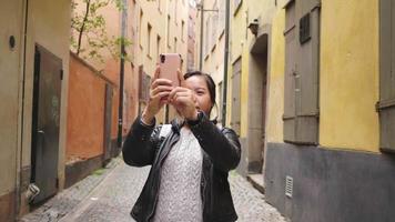 mujer asiática de pie y tomando una foto de un edificio colorido, usando un teléfono inteligente tomando una foto en una calle pequeña, un edificio colorido en el concepto de Suecia video