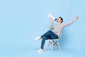 Portrait of young handsome Caucasian man sitting and cheerfully announcing on megaphone with one arm raised in isolated studio blue background photo