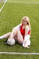 la rubia sostiene el balón entre las piernas en el campo de fútbol.
