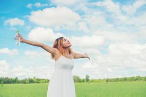 Women lifestyle concept young pretty  woman wearing white dress holding bottle of water at grassland smiley to camera stretching look so fresh enjoy and happy. photo