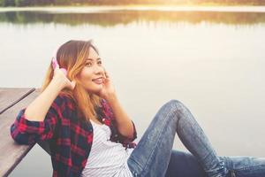 Portrait of young hipster woman with headphones sitting on pier and listening music relaxing with sunset,Lifestyle concept.
