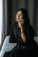 Woman wearing a white shirt, sitting on the bed and talking on the phone. photo