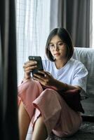 A woman wearing a white shirt, sitting on the bed and playing a smartphone. photo