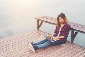 joven hipster feliz trabajando con su laptop sentada en el muelle, relajándose disfrutando con la naturaleza. foto