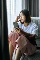 A woman wearing a white shirt, sitting on the bed and playing a smartphone. photo