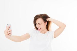 una mujer muy joven, una chica con camiseta se hace selfie en el teléfono inteligente con cámara frontal y muestra un pensamiento aislado en el fondo blanco. espacio en blanco, copia foto