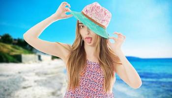 mujer joven y elegante muestra la lengua con vestido de color y sombrero de paja en la playa. foto