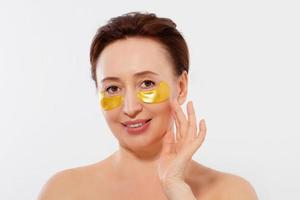 Macro female face. Beauty portrait of middle age woman with wrinkles and a gold patch under eye isolated on white background. Collagen mask and spa concept. Copy space. Summer skin care photo