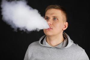 el hombre fuma un cigarrillo electrónico en el fondo oscuro, los hombres con capucha vapean y liberan una nube de vapor. chico con vaping sobre fondo negro foto