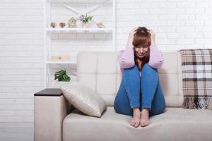 Sad middle aged woman sitting on sofa with clamped knees and crying. Home background photo