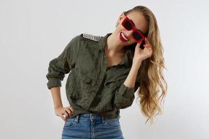 Stylish woman in red glasses looking at camera at studio isolated on white background. Copy space. Trendy style of beautiful luxury model with red lips makeup. Long wavy shiny blonde hair photo