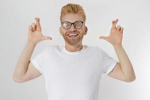 Fingers crossed, make a wish concept. Young redheaded guy with red beard cross finger for good luck. Hand sign, gesture for dreams come true. Happy smiling man in blank template white t shirt. Mockup photo