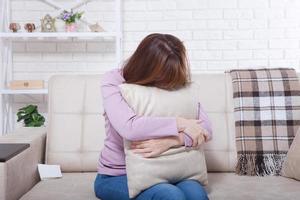 Beautiful middle aged brunette woman holding a pillow and crying on sofa. Home background, interior. Menopause time. Copy space photo