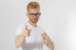 Guy pointing on you by fingers isolated on white background. Young stylish redheaded man, red beard and glasses. White t shirt shows on you. Success and confidence concept. Copy space. Selective focus photo