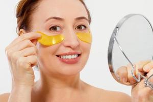 Wrinkle Macro female face. Beauty portrait of middle age woman with wrinkles and a gold patch under eye looking in mirror isolated on white background. Collagen mask and spa concept. Skin care photo