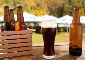 cup of dark beer and bottle on wooden table on blurred summer background photo