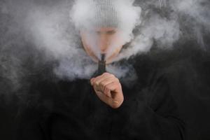 el hombre fuma un cigarrillo electrónico en el fondo oscuro, los hombres con capucha vapean y liberan una nube de vapor. chico con vaping sobre fondo negro foto