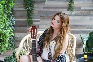 una hermosa chica con el pelo rizado rojo se sienta en una silla con una guitarra de siete cuerdas. foto