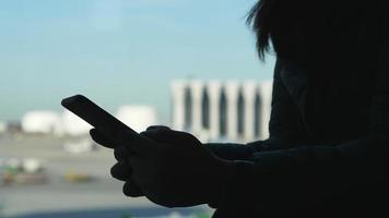 mani ravvicinate della donna in piedi e che utilizzano lo smartphone, digitando sullo smartphone, in piedi vicino alla finestra dell'aeroporto. mano ombra e modalità di visualizzazione luminosa video