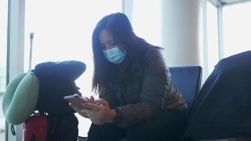 mulher asiática usando máscara, sentada e usando smartphone no aeroporto. esperando o voo no aeroporto com sua mala. indo para a cidade natal de férias de avião video