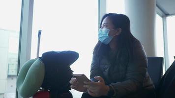 mulher asiática usando máscara, sentada e usando smartphone no aeroporto. esperando o voo no aeroporto com a mala, sentindo-se entediada e olhando pela janela. indo para a cidade natal de férias video