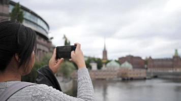 Back view of Asian woman standing and taking a picture of river and town in Sweden, traveling abroad on holiday. Using smartphone to take a photo. Beautiful town in Sweden video