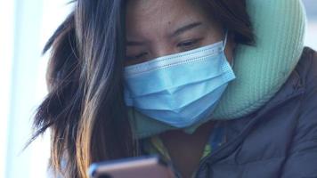 primer plano de una mujer asiática con máscara, sentada y usando un teléfono inteligente en el aeropuerto. esperando el vuelo en el aeropuerto con su maleta. ir a la ciudad natal de vacaciones en avión video