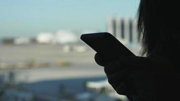 Close up hands of woman standing and using smartphone, typing on smartphone, standing by the window at the airport. Shadow hand and bright view mode video