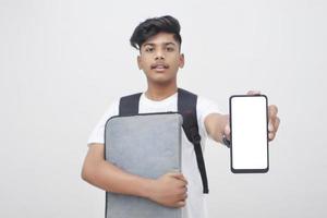 joven estudiante indio sosteniendo un archivo y mostrando la pantalla del teléfono inteligente con fondo blanco. foto