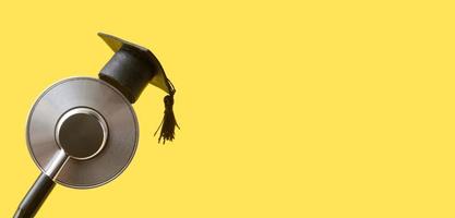 Graduation hat on medical stethoscope, yellow background with copy space banner format. Medical school, health care education or doctor's university degree concept photo