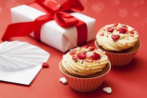 primer plano de dos cupcakes con decoración de crema y corazón sobre un fondo rojo con regalo y sobre.