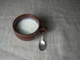 Plain white homemade yogurt in a brown clay cup, next to it is a silver spoon on a gray linen tablecloth. Minimalistic food background photo